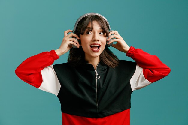 Excited young caucasian woman wearing headphones looking at side while grabbing headphones and listening to music isolated on blue background