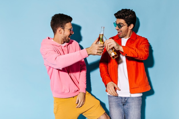 Возбужденные молодые очаровательные друзья в красочных солнцезащитных очках чокаются пивными бутылками Брюнет в оранжевой куртке и парень в розовой толстовке веселятся на синем фоне