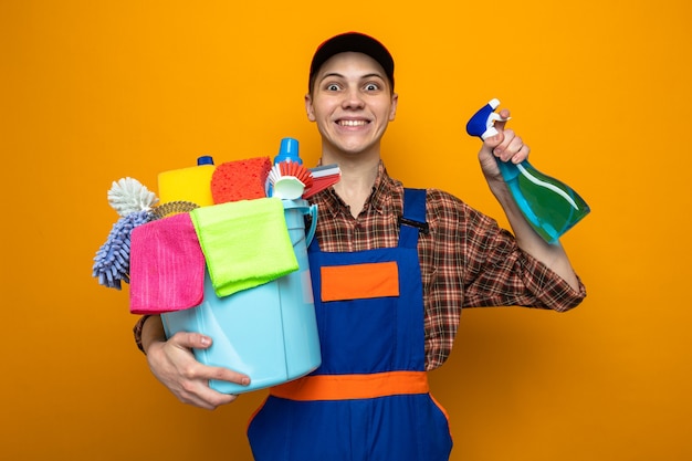 Excited young cleaning guy wearing uniform and cap holding bucket of cleaning tools with cleaning agent 