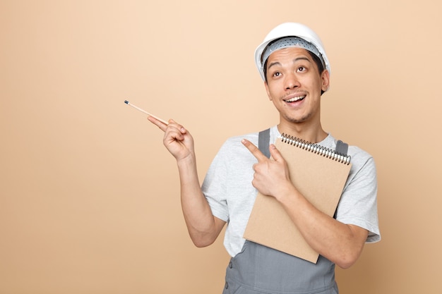 Excited young construction worker wearing safety helmet and uniform holding notepad and pencil looking up pointing up at corner 