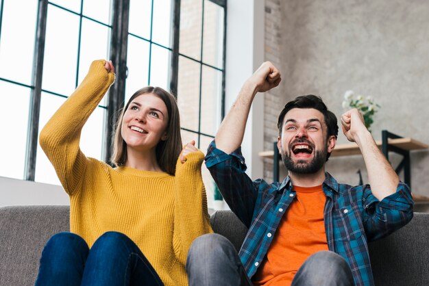 Excited young couple sitting on sofa cheering with joy at home