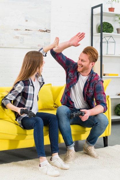 Excited young couple sitting on sofa holding joystick in hand giving high-five to each other