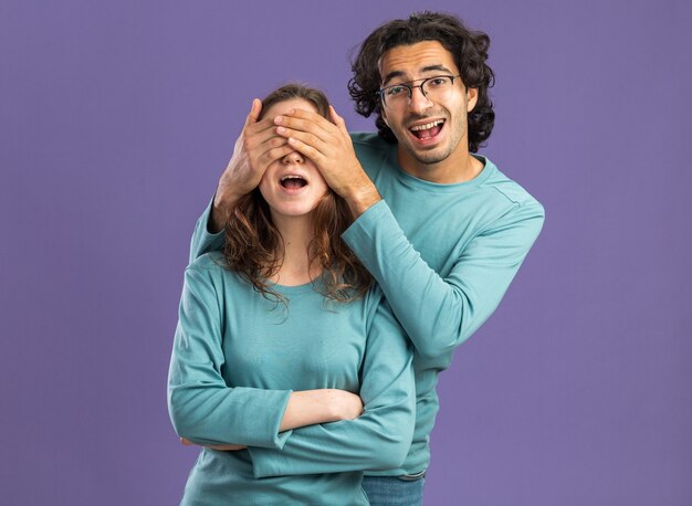 Excited young couple wearing pajamas man wearing glasses standing behind woman covering her eyes with hands looking at front woman standing with closed posture isolated on purple wall