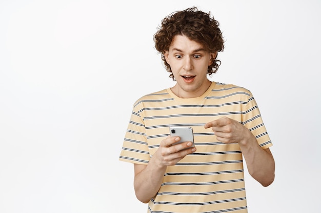 Возбужденный молодой кудрявый мужчина указывает и смотрит на свой телефон с удивленным улыбающимся лицом, проверяя что-то на смартфоне, стоящем на белом фоне
