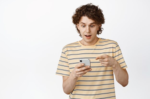 Excited young curly man pointing and looking at his phone with amazed smiling face checking out smth on smartphone standing over white background