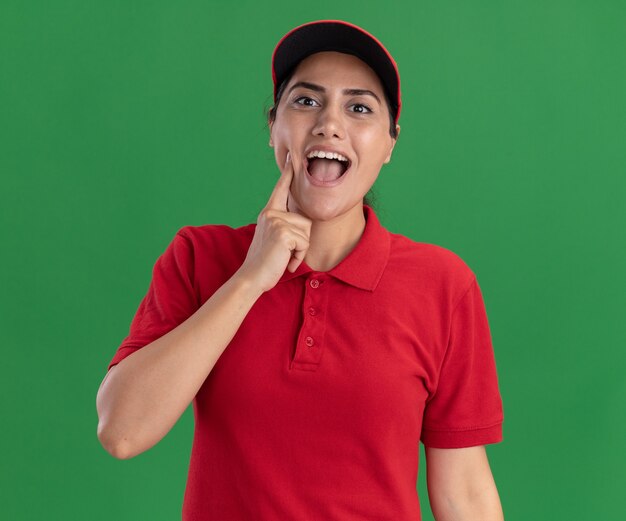 Возбужденная молодая доставщица в униформе и кепке положила палец на щеку, изолированную на зеленой стене