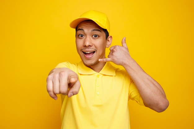 Excited young delivery man wearing cap and uniform looking and pointing at camera doing call gesture 