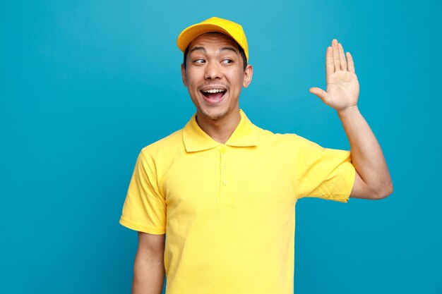 Excited young delivery man wearing uniform and cap looking at side doing hi gesture 