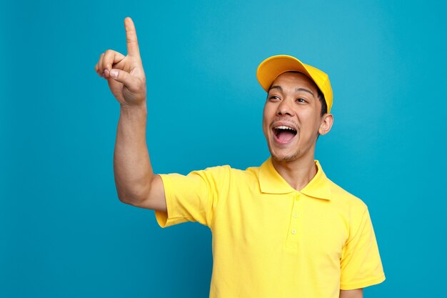 Excited young delivery man wearing uniform and cap looking up at corner pointing up 