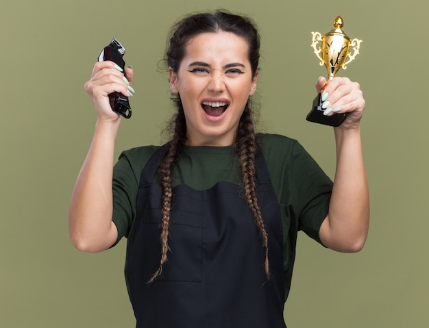 Excited young female barber in uniform holding hair clippers with winner cup isolated on olive green wall