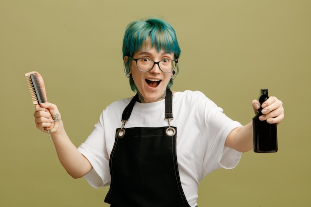 Excited young female barber wearing glasses uniform holding combs looking at camera stretching hair spray out towards camera isolated on olive green background