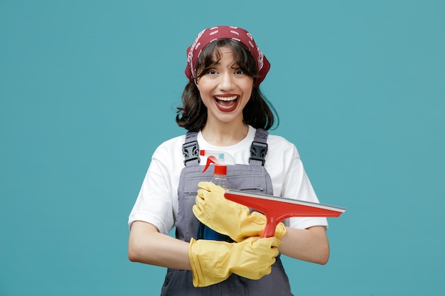 파란색 배경에 격리된 카메라를 바라보는 와이퍼와 클렌저를 들고 균일한 반다나와 고무 장갑을 끼고 흥분한 젊은 여성 청소부