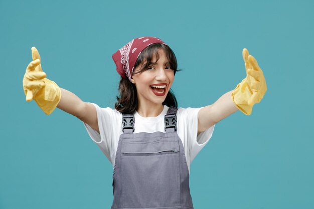 制服のバンダナとゴム手袋を身に着けている興奮した若い女性掃除機は、青の背景に両手を大きく広げて歓迎のジェスチャーを示すカメラを見て