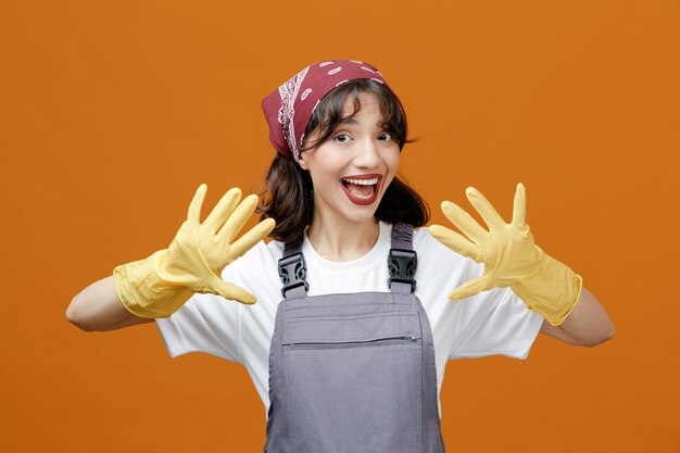 고무 장갑을 끼고 주황색 배경에 격리된 빈 손을 보여주는 카메라를 보고 있는 반다나를 착용한 흥분한 젊은 여성 청소부