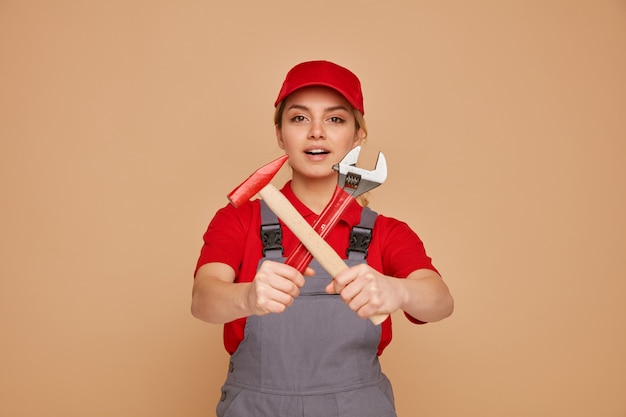 Excited young female construction worker wearing cap and uniform stretching out wrench and hammer towards camera doing no gesture 