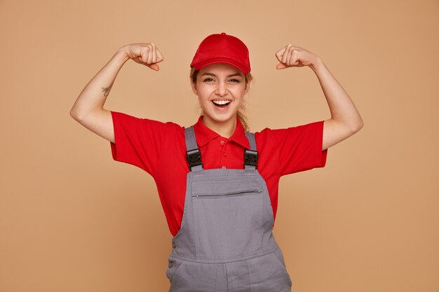制服とキャップを身に着けている興奮した若い女性の建設労働者は強いジェスチャーをしています