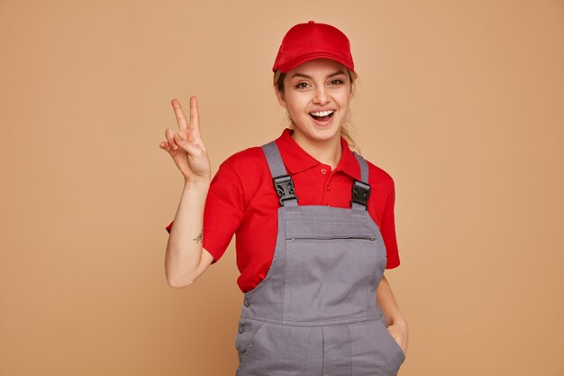 Excited young female construction worker wearing uniform and cap keeping hand in pocket doing peace sign