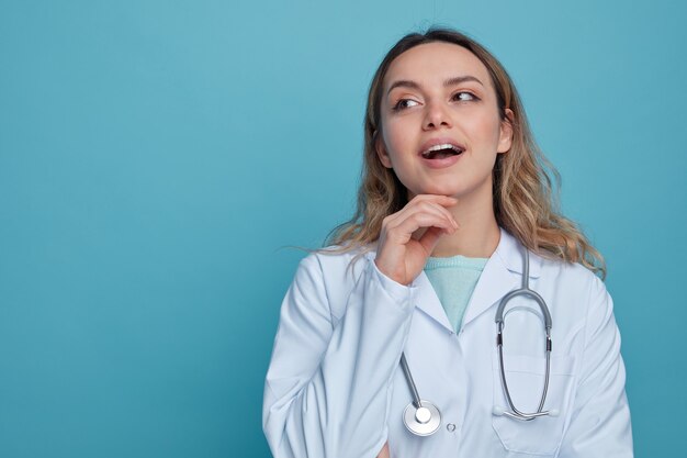 Excited young female doctor wearing medical robe and stethoscope around neck touching chin looking at side 