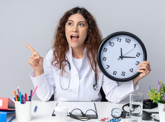 Возбужденная молодая женщина-врач в медицинском халате и стетоскопе сидит за столом с медицинскими инструментами, держит часы и смотрит в сторону, изолированную на белой стене