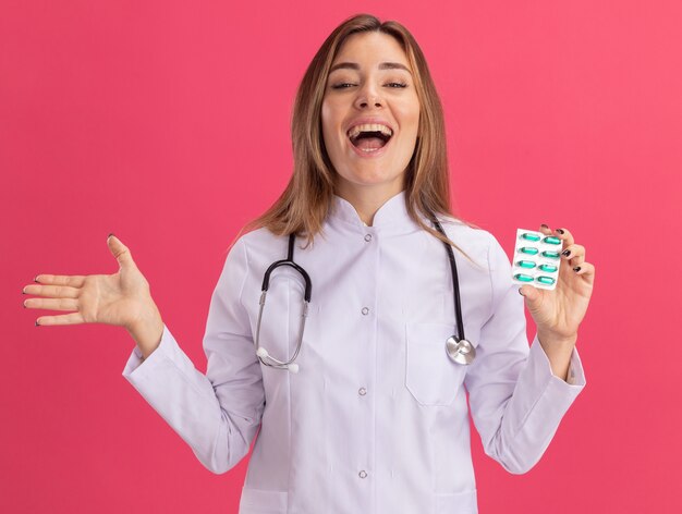 Excited young female doctor wearing medical robe with stethoscope holding pills spreading hand isolated on pink wall