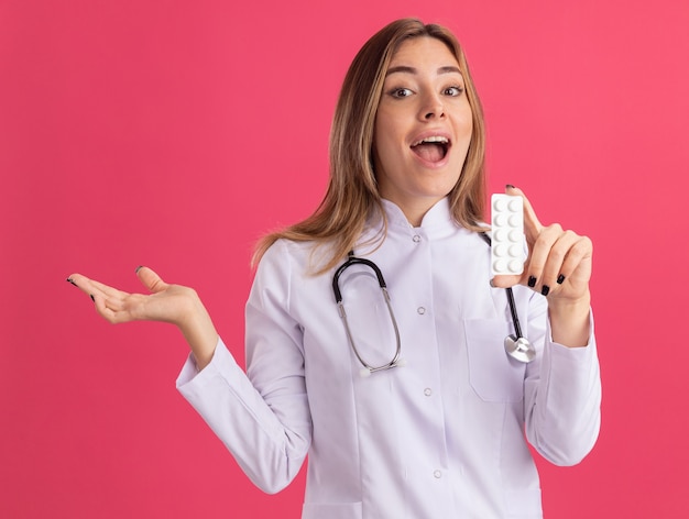 Eccitato giovane medico femminile che indossa abito medico con lo stetoscopio che tiene le pillole che diffondono la mano isolata sulla parete rosa