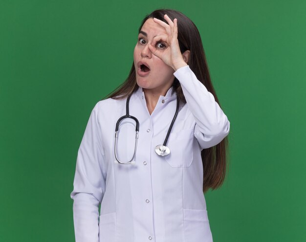 Excited young female doctor wearing medical robe with stethoscope looks at camera through fingers on green 