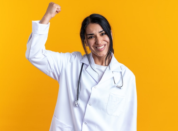 黄色の壁に分離された握りこぶしを上げる聴診器で医療ローブを身に着けている興奮した若い女性医師