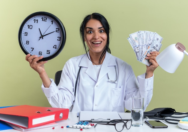 excited young female doctor wearing medical robe with stethoscope sits at desk with medical tools holding wall clock with cash isolated on olive green wall
