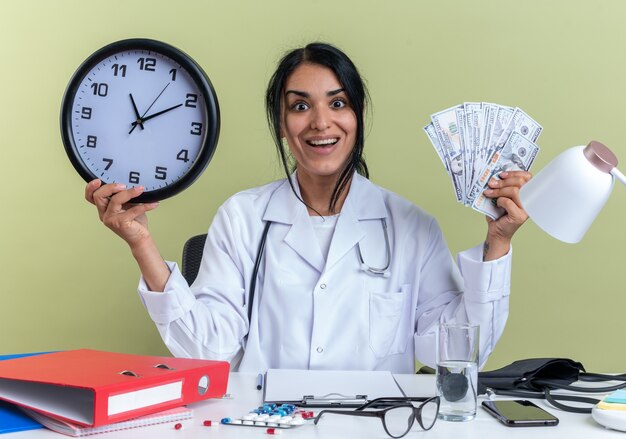 청진 기 의료 가운을 입고 흥분된 젊은 여성 의사는 올리브 녹색 벽에 고립 된 현금으로 벽 시계를 들고 의료 도구와 책상에 앉아
