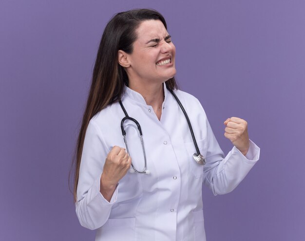 Excited young female doctor wearing medical robe with stethoscope stands with closed eyes keeping fists