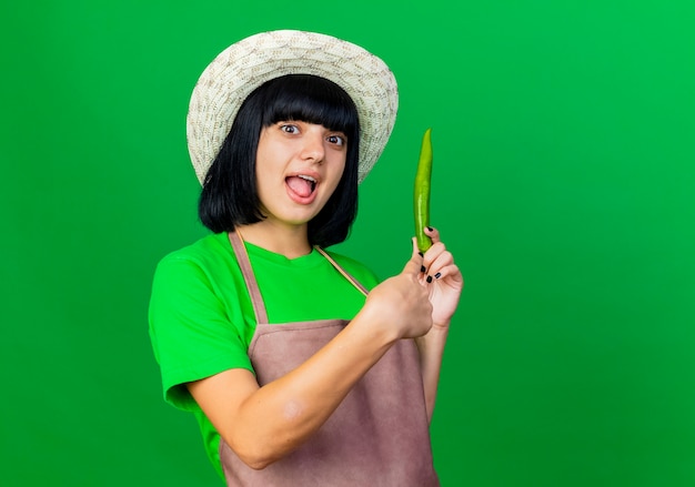 Возбужденная молодая женщина-садовник в униформе в садовой шляпе держит в изоляции острый перец