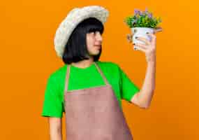 Free photo excited young female gardener in uniform wearing gardening hat holds flowerpot