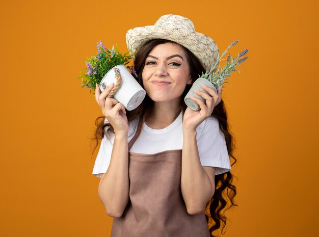 Excited young female gardener in uniform wearing gardening hat holds flowerpots isolated on orange wall with copy space