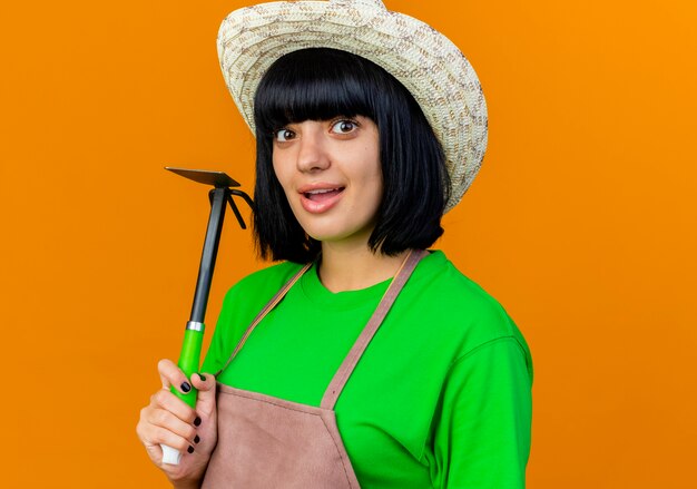 Excited young female gardener in uniform wearing gardening hat holds hoe rake