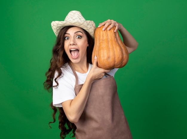 Il giovane giardiniere femminile emozionante in cappello da giardinaggio d'uso uniforme tiene la zucca isolata sulla parete verde con lo spazio della copia