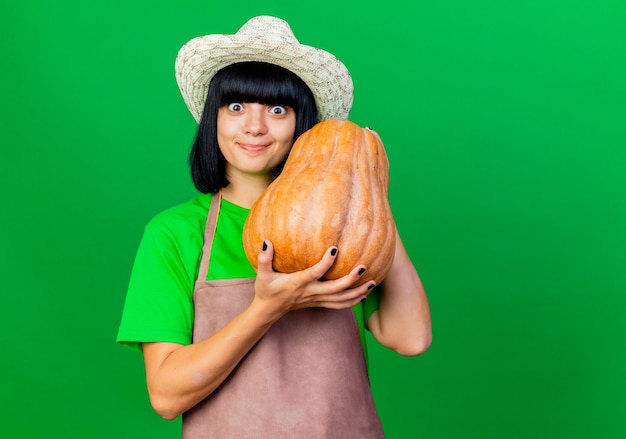 Excited young female gardener in uniform wearing gardening hat holds pumpkin 