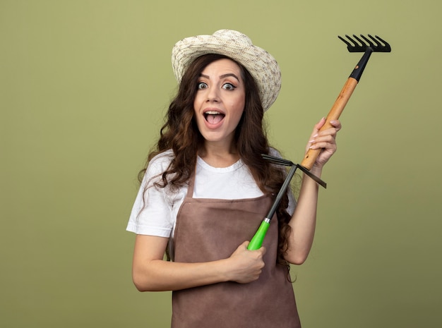 Excited young female gardener in uniform wearing gardening hat holds rake and hoe rake isolated on olive green wall