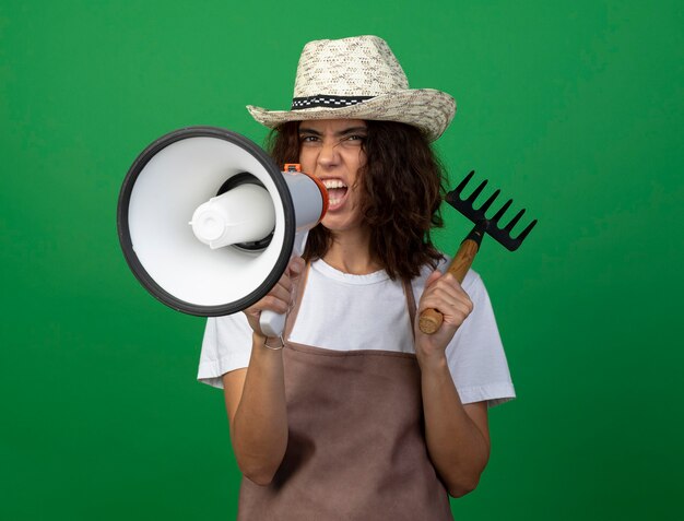 Eccitato giovane giardiniere femminile in uniforme che indossa cappello da giardinaggio mettendo il rastrello sulla spalla e parla sull'altoparlante