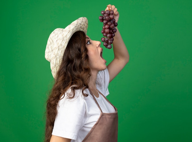 Excited young female gardener in uniform wearing gardening hat stands sideways pretending to bite grapes isolated on green wall