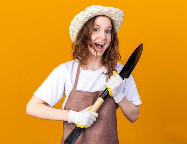 オレンジ色の壁に分離されたスペードを保持している手袋とガーデニング帽子をかぶって興奮した若い女性の庭師