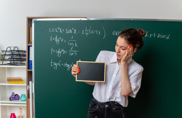 excited young female math teacher standing in front of chalkboard showing and looking at mini blackboard keeping hand on face in classroom