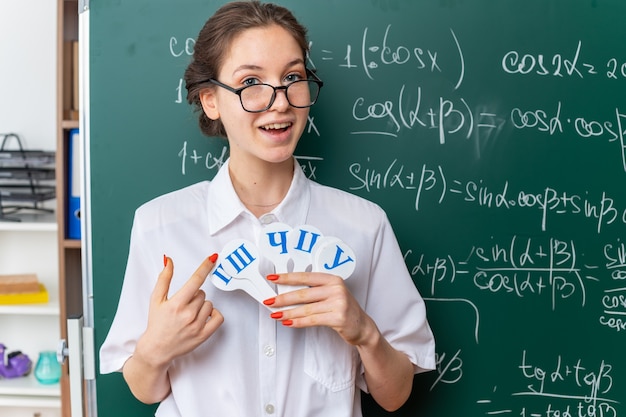 免费照片兴奋的年轻女数学老师戴眼镜站在黑板前持有和俄文字母表指着信球迷看在教室前面