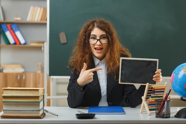 안경을 쓰고 교실에 학교 도구를 들고 책상에 앉아 있는 미니 칠판을 가리키는 흥분한 젊은 여교사