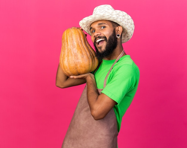 Excited young gardener afro-american guy wearing gardening hat holding pumpkin