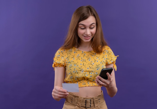 Excited young girl holding mobile phone and card and looking at mobile phone on isolated purple space with copy space