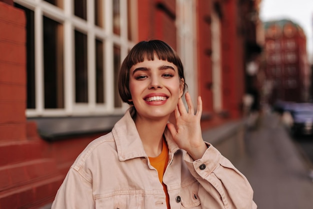 Excited young girl in pink denim jacket smiles sincerely outdoors Happy pretty shorthaired woman walks outside