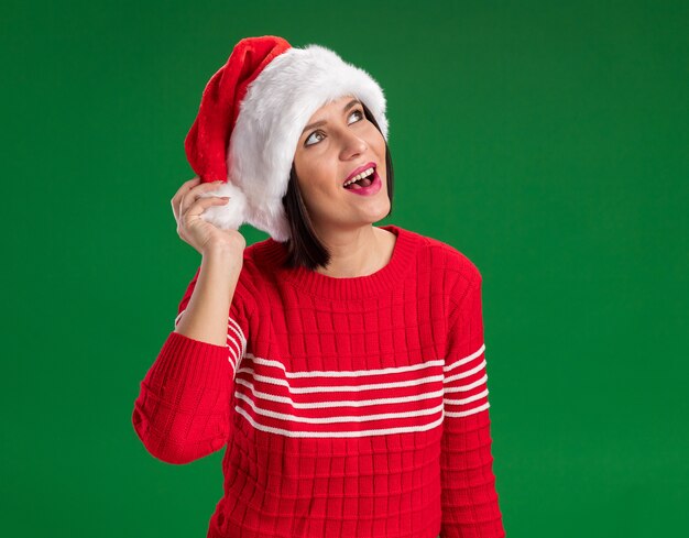 Excited young girl wearing santa hat grabbing hat looking up isolated on green background with copy space