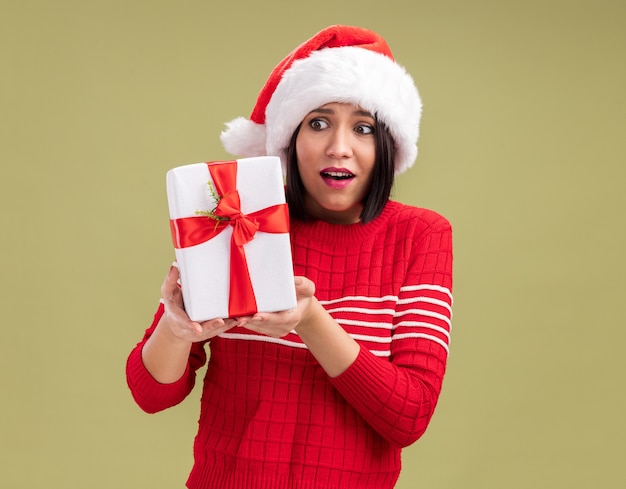 Excited young girl wearing santa hat holding and looking at gift package isolated on olive green background with copy space