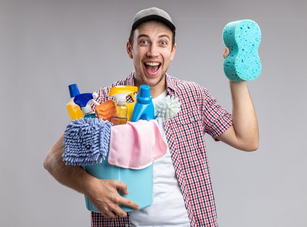 Excited young guy cleaner wearing cap holding bucket with cleaning tools and cleaning sponge isolated on white background