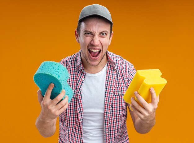 Excited young guy cleaner wearing cap holding cleaning sponges isolated on orange background